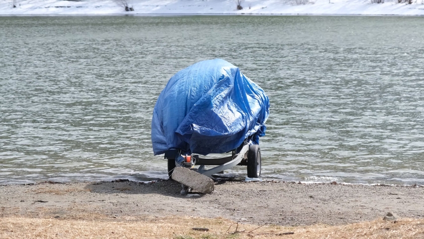 modern motorboat ship covered with blue protection tarp standing on the shore of the lake. Boat vessel on the background of a snow-covered shore