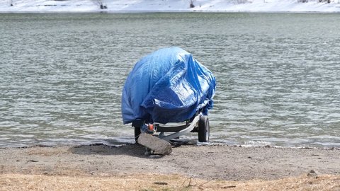 modern motorboat ship covered with blue protection tarp standing on the shore of the lake. Boat vessel on the background of a snow-covered shore