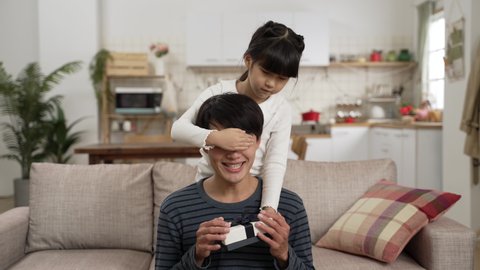 asian girl covering father’s eyes and giving him a gift box from behind on father's day at home. she asks her dad to guess what is inside as the man is holding and feeling the present