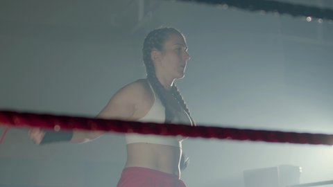 Young Caucasian woman with pigtails jumping rope in boxing ring while warming up before training in boxing gym. Female boxer with bandages on fists exercising before fight. Sport, exercising concept