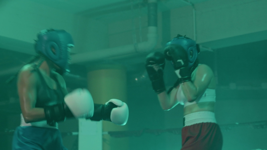 Two female boxers in helmets and boxing gloves fighting in ring at gym. Young sportswoman hitting sparring partner on chin while girls training and practicing self-defense. Sport, boxing concept
