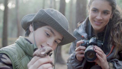 Happy mother and son exploring wildlife in natural park. Smiling woman taking photo of boy holding mushroom and posing at camera. Close-up shot. Leisure, nature concept