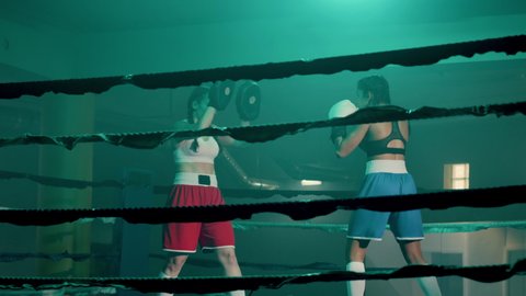 Two female boxers training in boxing gym together. Slim athletic girl in blue shorts practicing punches with boxing gloves while sparring partner holding boxing paws. Sport, fighting, box concept