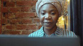 Beautiful African American woman in head wrap waving and talking on online video call on laptop while sitting in cafe