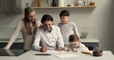 Happy family parents and two diverse children older brother younger sister having fun talk engaged in drawing at home kitchen. Smiling mom preteen boy watch dad little girl coloring pictures in album