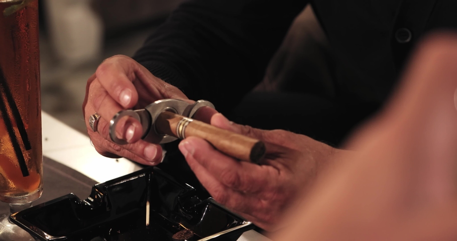 Male hands with a gold ring on their finger are cutting a cigar with a guillotine of scissors, on a table near a cocktail over an ashtray. close-up | Shutterstock HD Video #1089230991
