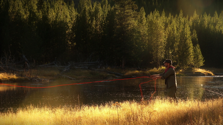 Fly Fisherman casting his line in scenic wild river in slow motion backlight. Golden hour light in rocky mountains of Yellowstone National Park, Wyoming and Montana, USA. 4K.