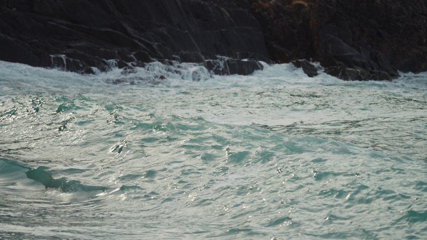 S close-up shot of the waves crashing on the sandy beach. Black rocks in the background. Slow-motion.
