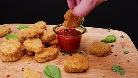 Dipping vegetarian vegan chicken nuggets in sauce on a bamboo cutting board with basil leaves and peppercorns for garnish, black background, static shot