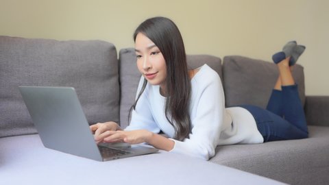A young woman lying on her stomach on the couch works successfully on her laptop.
