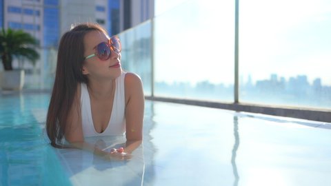 Close-up of a pretty woman lying on her stomach in the rooftop swimming pool as she looks out over a modern city skyline.