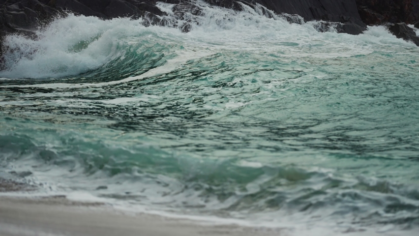 Powerful waves are crashing on the rocks and spilling on the sandy beach. Slow-motion, close-up shot.