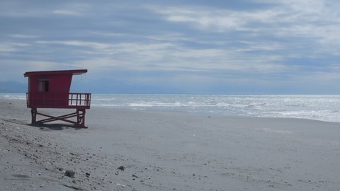 Male creator steps on sandy beach, throws backpack and shoots vertical photo. Old vintage wooden baywatch tower on background, empty coast, calm see, blue sky and clouds. Slow motion footage.