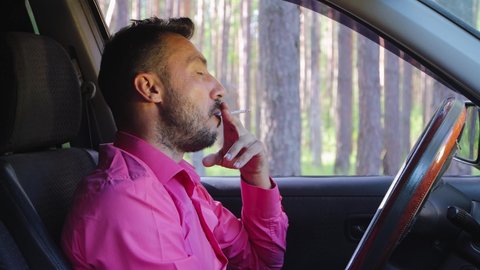 Serious businessman smoking a cigarette while driving and talking. A young man in a car on a background of green trees, a hot sunny summer day. Warm soft light, close-up. 4K UHD.