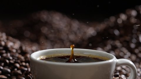 Drop of coffee falling into coffee cup sitting in a pile of freshly roasted coffee beans. Slow motion.