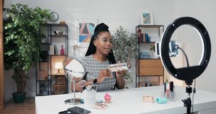 Young beauty influencer shoots video for her social media channel using phone and ring light. The makeup artist shows eyeshadow palette, talks about oigmentation, blending colors gestures with brush.