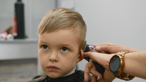 Hairdresser girl cuts the hair of blonde child with an electric trimmer machine. Process cutting hair in barber shop young girl. Men haircut and hair styling in salon. Cutting hair with trimmer.