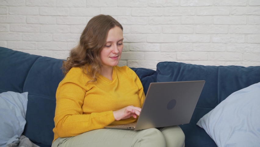 Woman is sitting on the sofa with laptop and typing something. Correspondence with friends, she read something and got scared, shock on her face. Work at home, e-learning, quarantine, distance