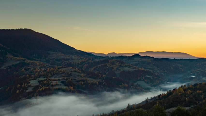 Sun rises in highland with big forestry mountains and morning fog fills valley among hill slopes, autumn under orange sky 4K time-lapse video