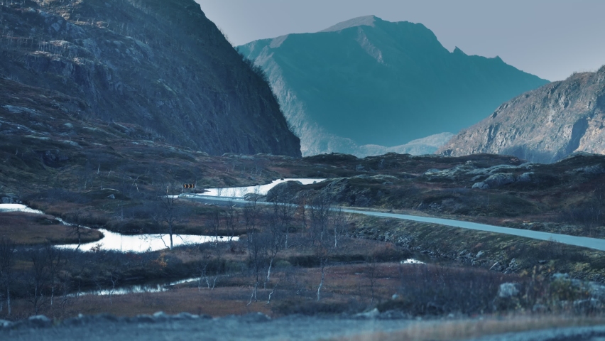 A narrow road snaking through the tundra. Mountains tower on both sides. lakes and puddles scattered through the valley. Aerial view.