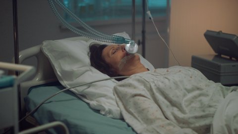 Oxygen mask patient lying in empty hospital ward. Medical tools table closeup. Unconscious aged woman in coma attached to artificial breathing tube lying in clinic bed. Terminally ill person concept.