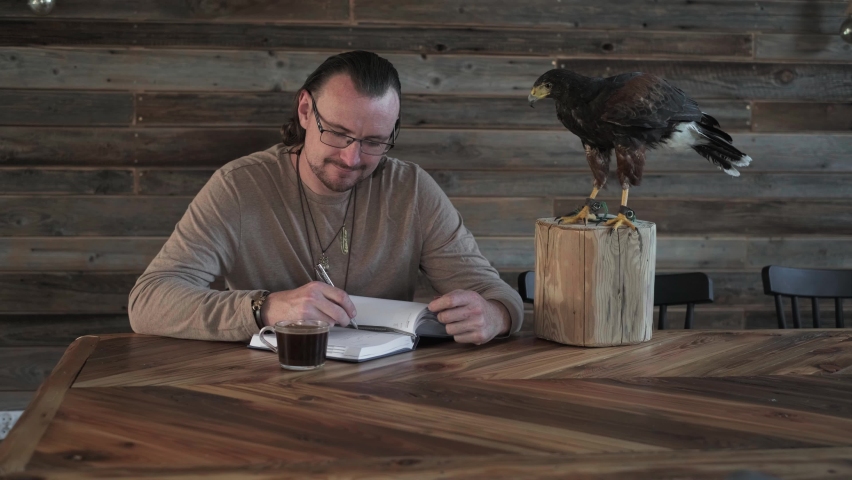 Man is working, writing with wild bird at home by the table. Making noted, memories, diary with eagle as pet. Unusual animals at home. Human friendship. Taking care of buzzard