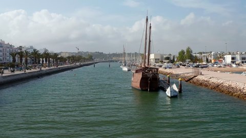 Portuguese wooden caravela moored in harbor town of Lagos Algarve. Aerial drone flying forward towards sailing boat in port canal, day in 2021