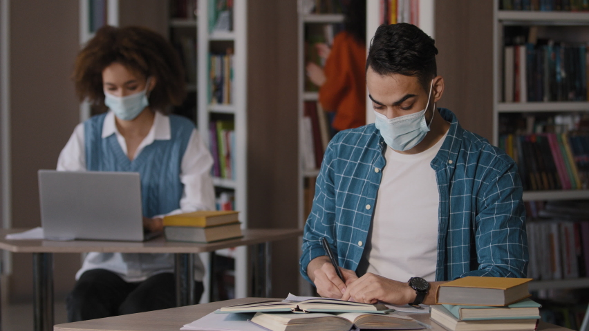 Tired overworked young arab male student in protective mask sits at desk in library does homework writes notes prepares for exam makes mistake grabs head with hands feels shock frustration failure Royalty-Free Stock Footage #1089273505