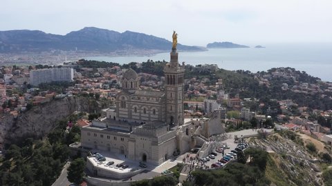 France, Marseille. Drone aerial view around Notre-Dame de la Garde basilica with its gold statue looking the mediterranean sea, on a sunny day with blue sky. Calanques mountain in back.