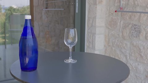 Close up view of person's hand pouring mineral water from blue bottle into glass on patio in hotel. Greece.