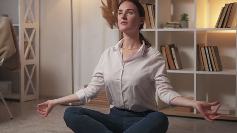 Home yoga. Weekend meditation. Stress relief. Relaxed woman sitting cross-legged in lotus position practicing breathing technique on floor at cozy living room interior.