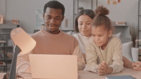 Medium slowmo of young happy interracial couple and their cute 8 year old daughter watching funny videos on laptop, staying at home together during lockdown