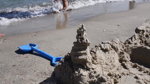 Sand castle and blue plastic toy shovel on beach sand and barefoot feet of people walking on background. Sand shovel in sea surf, waves wash seashore. Vacation beach summer game sand construction