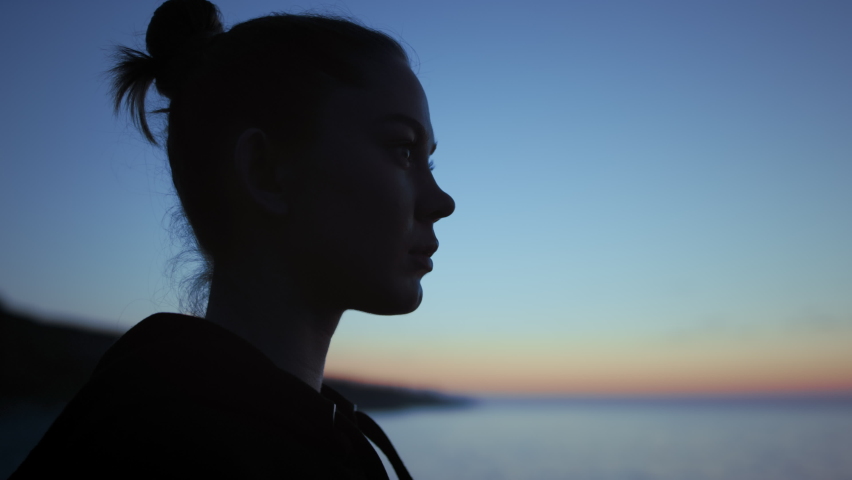Dark silhouette face woman standing in front blue evening sky close up. Calm girl relaxing on beach summer twilight. Unrecognizable lady watching picturesque waterscape outdoors. Nature concept.