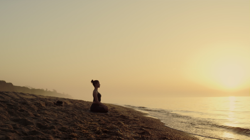 Focused woman practicing meditation on sandy beach summer evening. Yoga woman sitting lotus position near ocean waves. Attractive sportswoman making relaxing asana outdoors. Self care concept.