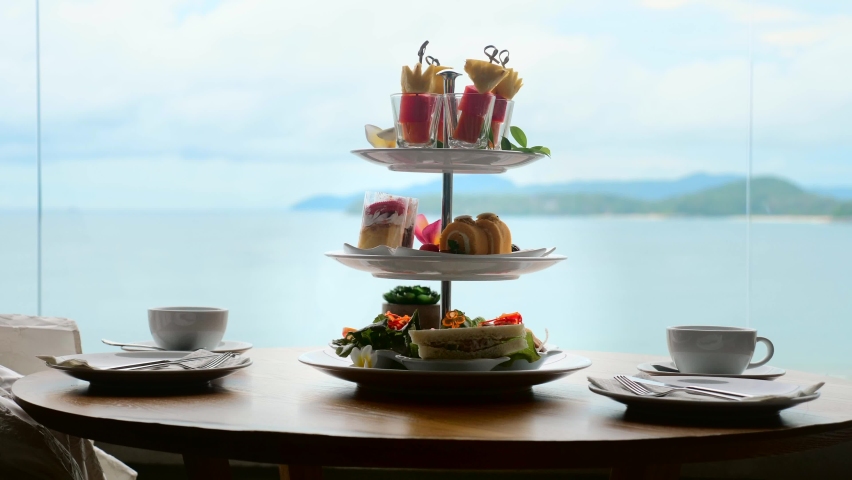 Woman hand take skewer with fruit slices from stand with cakes and sweet pastries in cafe with sea view. Afternoon tea in restaurant. Romantic, elegant, high class tea.