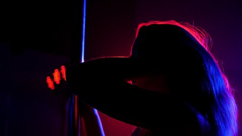 Close-up silhouette of a sexy woman with long flowing hair near a pole on a background of bright colored spotlights in a nightclub. Pole dance.