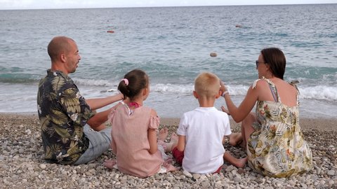 A happy family is sitting on the beach on the seashore in the evening on small stones, they hug together and smile. View from the back.