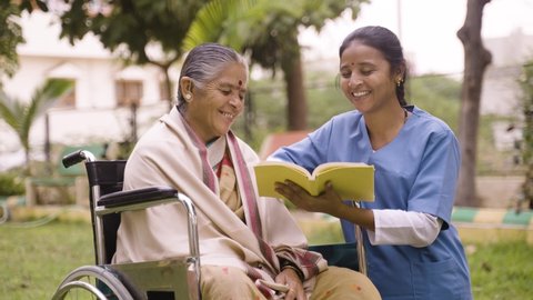 Nurse and senior woman on wheel chair laughing by reading book or novel at park - concept of relaxation, professional occupation, therapy or treatment – Stockvideo
