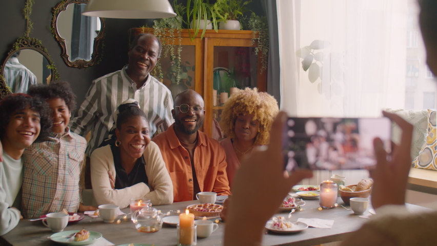 Large African American family smiling and posing together on camera at home holiday dinner while kid taking picture with smartphone