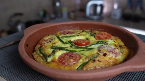 Ceramic bowl with vegetable frittata, simple vegetarian food. Frittata with egg, tomato, pepper, onion, cheese and green wild garlic leaves on the table, rotate, close up. Healthy egg omelet