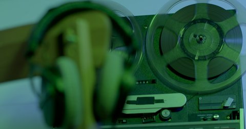 The movement of reels of tape on a tape recorder in green light. Using an old recorder to record and listen to audio. Headphones on the stele in bokeh.