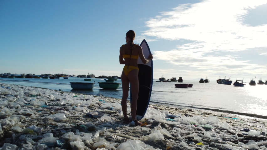 Caucasian young athletic female surfer stand in polluted beach with plastic waste trash garbage, Mother Earth ocean pollution save the planet concept