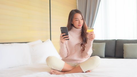 A young, attractive woman sits cross-legged on the bed as she inputs her credit card number into her smartphone. Title space