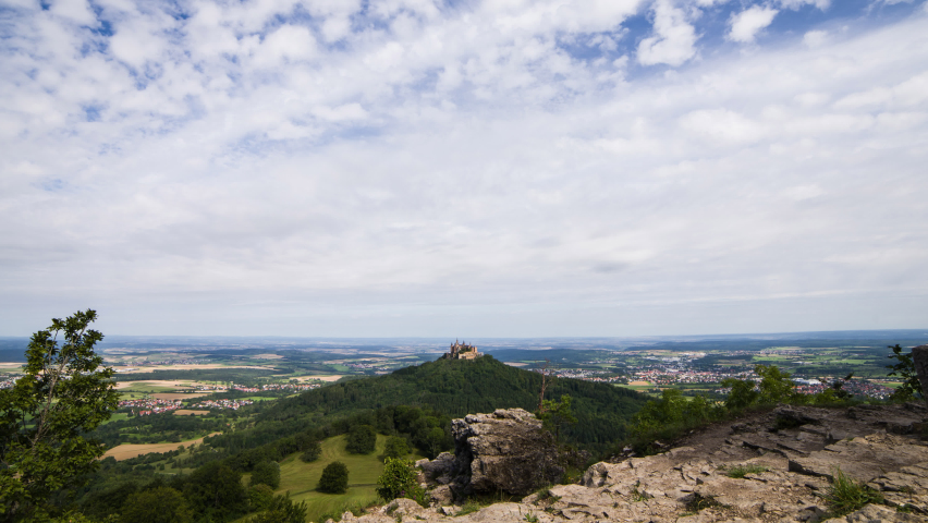 Timelapse image of clouds in the landscape, with Hohenzollern Castle (Swabian Alb, Germany). The hill towers above the residential areas in the valley. The sky is blue and the meadows are green.