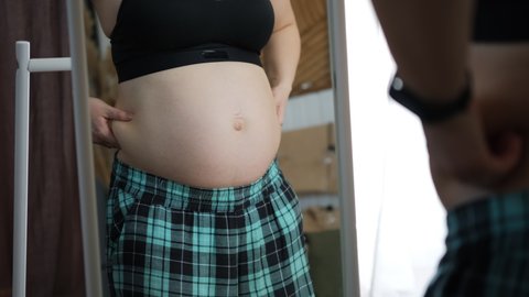 Pregnant Woman Pinching her Potbelly near Mirror Indoors, Unhappy to See her Weight Increasing. Expectation of a Baby Concept