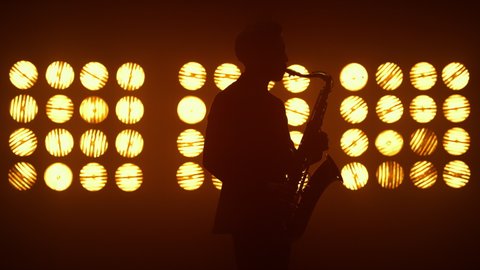 Unknown jazz man saxophonist performing on stage lighted spotlights. Silhouette of musician playing music professionally on scene nightclub. Talented player holding modern sax on concert indoor.