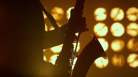 Unknown saxophonist hands playing musical instrument solo in studio spotlights close up. Professional saxophone player performing on stage night club. Silhouette of man musician holding sax indoors.