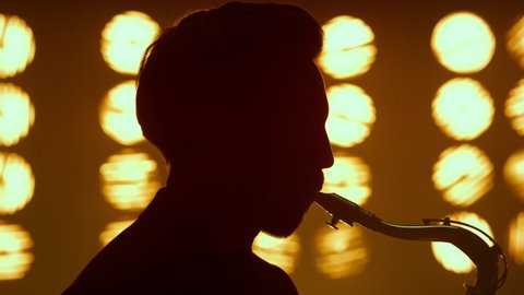 Dark silhouette of guy blowing in saxophone standing nightclub stage closeup. Portrait of unknown man playing musical instrument in night club spotlights. Young saxophonist performing in studio lights