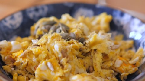 Close up view of Scrambled eggs with sunflower seeds and nuts in a blue bowl on a brown kitchen table